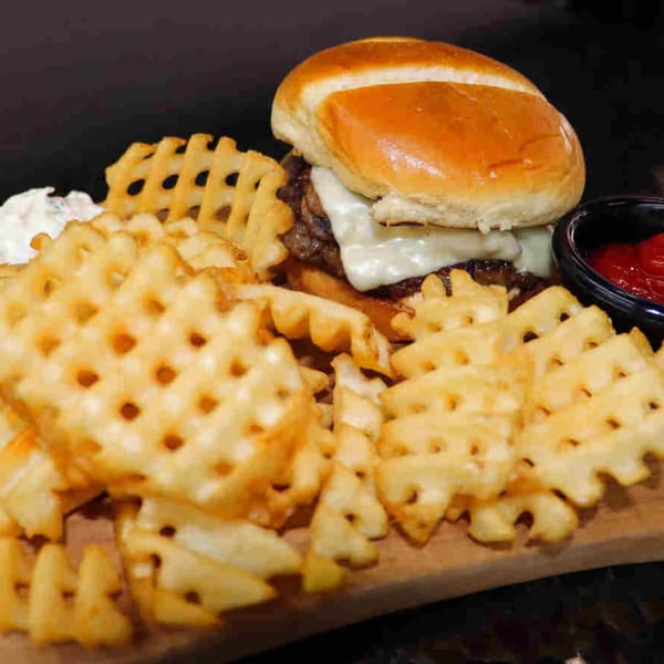Cheeseburger with melted white cheese, served with waffle fries, white sauce, and ketchup on a wooden board.